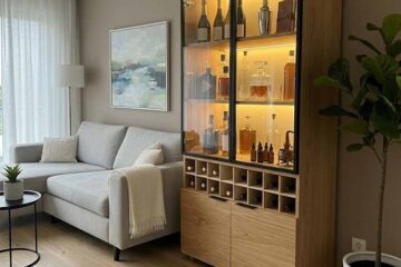 Living room with a tall wooden bottle cabinet lit from inside, beside a gray sofa and a potted plant.