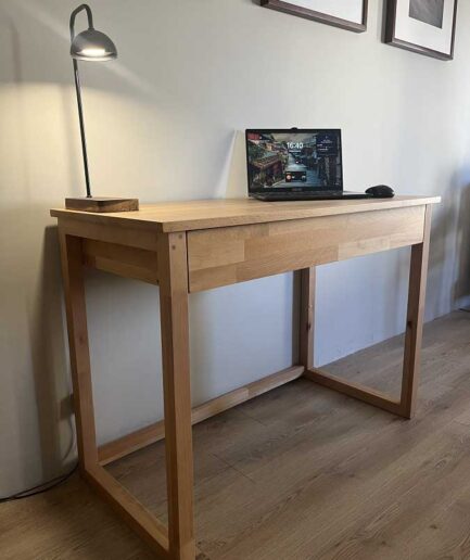 Wooden desk with a laptop and a black desk lamp on top, against a pale wall with framed photos.