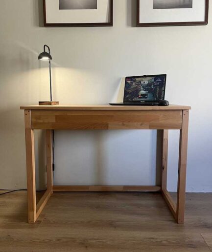 Wooden desk with a laptop, a small lamp on the left, and two framed photos on the wall above in a simple home office.